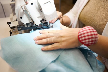 Téléchargez les photos : Femme travaille sur une machine à coudre avec du tissu bleu, elle a un coussin d'épingle sur la main - en image libre de droit