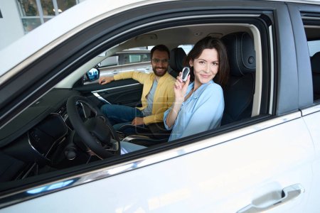 Foto de Hermosa mujer sosteniendo la llave del coche y mirando a la cámara mientras se sienta en auto con su novio en el centro del coche - Imagen libre de derechos