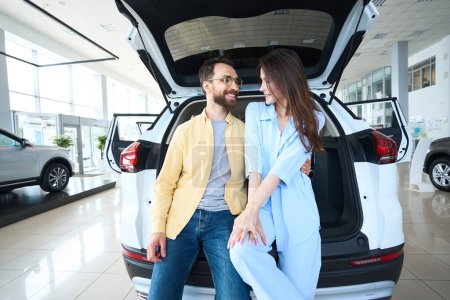 Téléchargez les photos : Femme caucasienne et homme appuyé sur le coffre de la voiture tout en se regardant dans la salle d'exposition de la voiture - en image libre de droit
