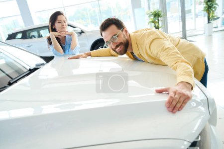 Foto de Hombre guapo abrazando capucha de auto mientras mira a su novia mirándolo en concesionario de automóviles - Imagen libre de derechos