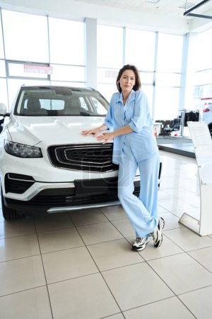 Photo for Attractive woman in stylish clothes looking at camera while leaning on auto in car dealership - Royalty Free Image