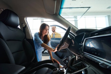 Foto de Mujer feliz sosteniendo el volante del coche mientras su novio de pie detrás de ella en la concesionaria de automóviles - Imagen libre de derechos