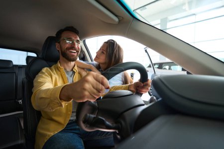 Foto de Sonriente hombre sentado en el interior del coche y sosteniendo el volante mientras su novia coquetea con él en el centro del coche - Imagen libre de derechos
