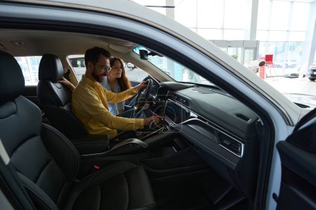 Foto de Hombre guapo sentado en el interior del coche y sosteniendo el volante mientras su novia lo observa en el concesionario de automóviles - Imagen libre de derechos