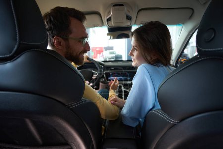 Téléchargez les photos : Homme souriant dans des lunettes et femme caucasienne se regardant tout en étant assis à l'intérieur de l'automobile dans le concessionnaire automobile - en image libre de droit