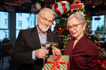 Photo for Aged woman and man clinking glasses of champagne holding gift box celebrating Christmas in restaurant - Royalty Free Image