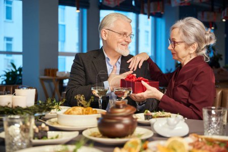 Photo for Mature couple celebrating New Year in restaurant man giving present box to woman - Royalty Free Image