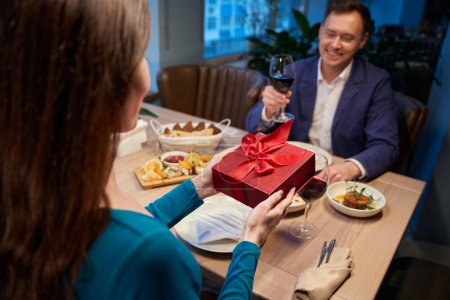 Photo for Man and woman having romantic dinner female giving red gift box making present during Christmas celebration in restaurant - Royalty Free Image