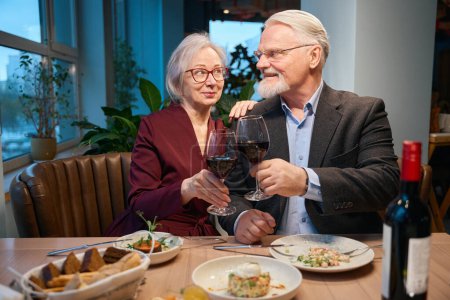 Foto de Linda pareja de edad tintineando gafas de vino celebrando la Navidad juntos disfrutando de la víspera de Año Nuevo - Imagen libre de derechos