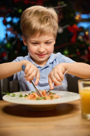 Foto de Niño pequeño que usa tenedor y cuchillo mientras cena en el restaurante durante la celebración de Navidad - Imagen libre de derechos