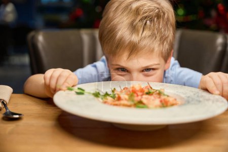 Téléchargez les photos : Petit garçon drôle regardant assiette avec plat de fête tout en célébrant le Nouvel An au restaurant - en image libre de droit