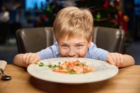 Foto de Alegre niño divertido mirando la comida del apetito en la mesa en el restaurante mientras se celebra la Navidad - Imagen libre de derechos