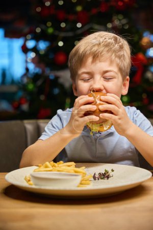 Foto de Niño hambriento disfrutando de una hamburguesa grande durante la celebración de Navidad en un acogedor café decorado - Imagen libre de derechos