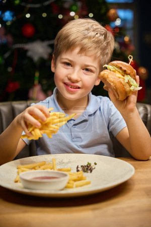 Téléchargez les photos : Petit garçon joyeux montrant un gros hamburger à la fête de Noël au restaurant célébrant le Nouvel An avec menu de restauration rapide - en image libre de droit