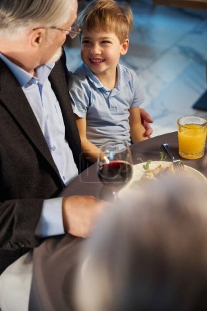 Téléchargez les photos : Grand-père et petit-fils assis à une table festive parlant et souriant dans un restaurant confortable - en image libre de droit
