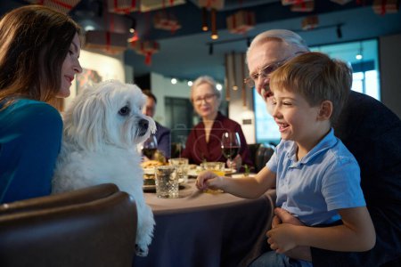 Foto de Mujer sonriente con perro en las manos celebrando el día de Acción de Gracias junto con la familia en el restaurante festivo - Imagen libre de derechos