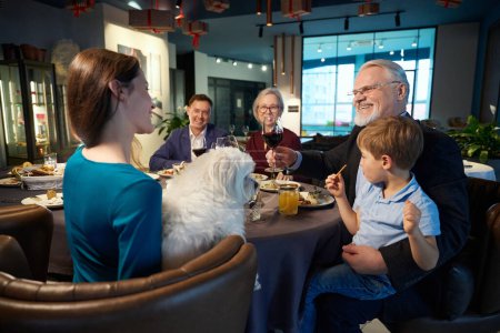 Photo for Smiling old man with glass of wine talking with family while celebrating Christmas in restaurant - Royalty Free Image
