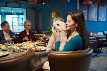 Photo for Beautiful smiling woman with dog in hands sitting at table in restaurant during family celebration of New Years Eve - Royalty Free Image