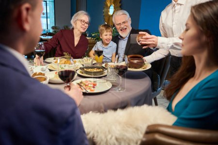 Photo for Happy old man making selfie with little boy and aged woman while sitting at table in restaurant - Royalty Free Image
