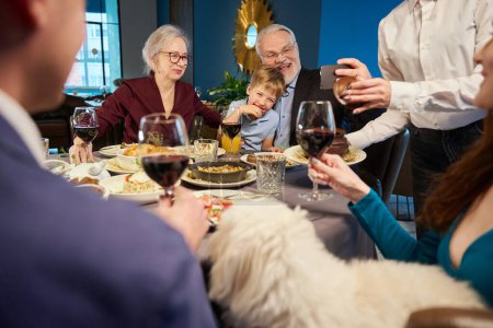 Téléchargez les photos : Homme extrêmement heureux passé du temps avec la famille faire du selfie avec smartphone tout en célébrant Noël au restaurant - en image libre de droit