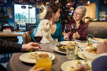 Photo for Smiling women with glasses in hands drinking red wine celebrating winter holidays together in cozy restaurant - Royalty Free Image