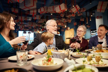 Téléchargez les photos : Joyeux vieil homme avec un verre de vin grillé saluant sa famille avec des vacances de Noël tout en étant assis ensemble dans un café - en image libre de droit
