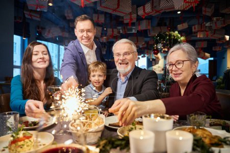 Photo for Cheerful family with sparkling Bengal lights enjoying Christmas party at cozy decorated restaurant - Royalty Free Image