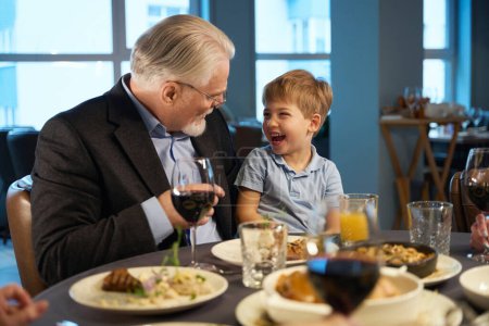 Téléchargez les photos : Grand-père et petit-fils assis à une table festive mangeant dans un restaurant confortable - en image libre de droit