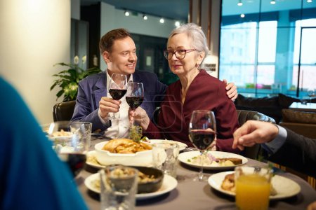 Photo for Man and old lady sitting at a table in a cozy restaurant clinking glasses of wine, celebrating new year - Royalty Free Image
