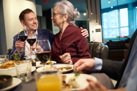 Téléchargez les photos : Homme adulte et vieille dame assis autour de la famille à une table festive cliquetis vin de verres et de manger - en image libre de droit