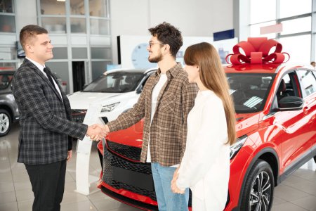 Téléchargez les photos : Homme et femme joyeux serrant la main de l'agent de vente après avoir réussi l'achat de voiture dans le concessionnaire - en image libre de droit