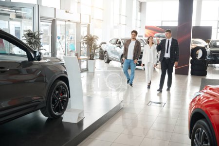 Photo for Young couple choosing new car waking around dealership with salesman choosing suitable model of auto - Royalty Free Image