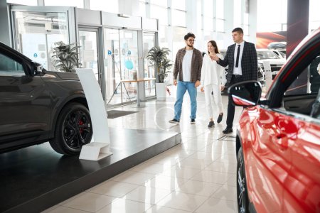 Foto de Atractivo hombre y mujer elegir y comprar un coche en la sala de exposición automática con vendedor ayuda a caminar alrededor de concesionario - Imagen libre de derechos