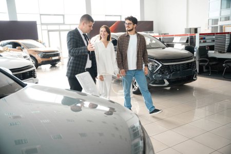 Foto de Pareja joven hablando con la persona de ventas en la sala de exposición de automóviles elegir el automóvil para el uso diario - Imagen libre de derechos