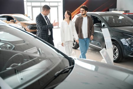Foto de Joven hombre de las ventas que habla con los clientes del hombre y de la mujer que ayudan a elegir gran automóvil en concesionario - Imagen libre de derechos