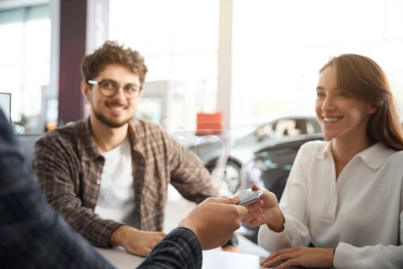 Téléchargez les photos : Un homme et une femme souriants assis dans un bureau de concession reçoivent les clés de leur nouvelle automobile - en image libre de droit