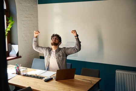 Photo for Young excited successful caucasian businessman celebrating win in work at desk in coworking office. Concept of modern business lifestyle - Royalty Free Image