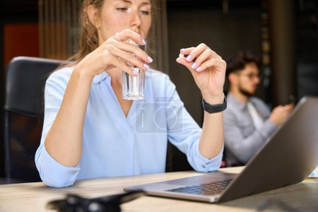 Photo for Selective focus of young sick caucasian businesswoman taking pill with water during work at desk with blurred male colleague on background in office. Concept of workaholism and working burnout - Royalty Free Image