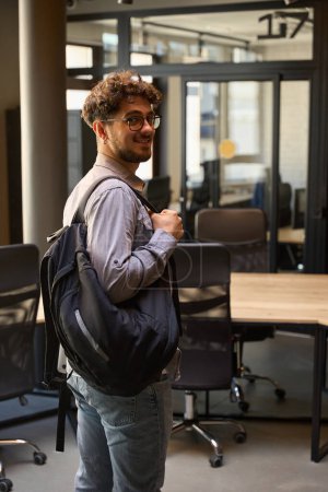 Photo for Young smiling caucasian businessman with backpack and wearing glasses standing and looking at camera in empty coworking office. Concept of modern business lifestyle - Royalty Free Image