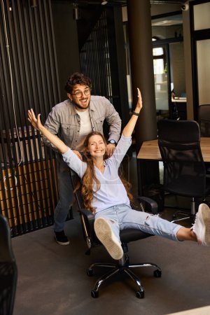 Photo for Excited businessman riding on office chair his young laughing female colleague with hands in air in coworking office. Concept of break and rest at work - Royalty Free Image