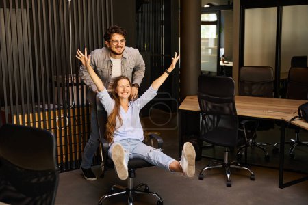 Téléchargez les photos : Homme d'affaires chevauchant sur chaise de bureau sa jeune collègue joyeuse avec les mains dans l'air pendant qu'ils plaisantent dans le bureau de coworking. Concept de pause et de repos au travail - en image libre de droit