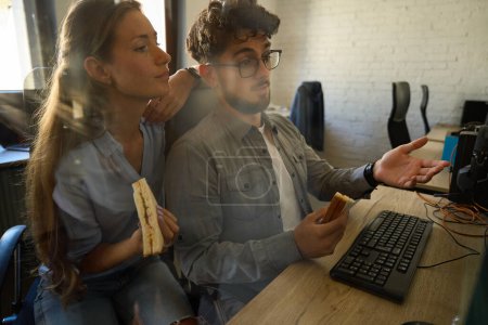 Photo for Dissatisfied businessman showing something on computer to focused female colleague during they eating sandwiches in coworking office. Concept of teamwork - Royalty Free Image