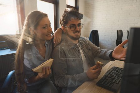 Téléchargez les photos : Homme d'affaires montrant quelque chose sur l'ordinateur à une collègue féminine concentrée pendant qu'ils sourient et mangent des sandwichs dans le bureau de coworking. Concept de travail d'équipe - en image libre de droit