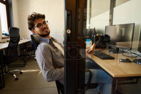Photo for Side view of smiling businessman eating sandwich on lunch and looking at camera during work at desk in coworking office. Concept of break and rest at work - Royalty Free Image