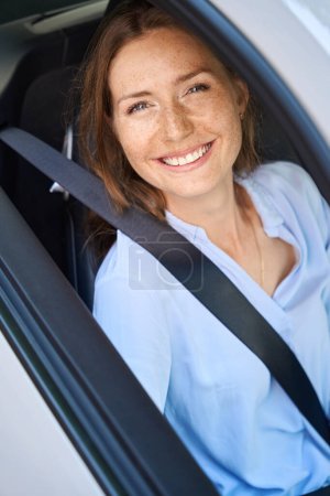 Téléchargez les photos : Jeune femme caucasienne souriante regardant la caméra par la fenêtre de la voiture. Concept de road trip, voyage et vacances - en image libre de droit