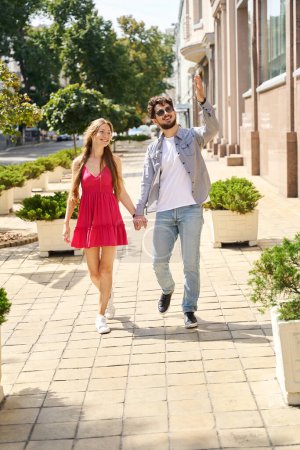 Photo for Young smiling caucasian man showing something to his pleased girlfriend while they walking on city pavement in warm summer day. Concept of romantic relationship and date - Royalty Free Image