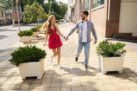 Photo for Young pleased caucasian couple holding hands, walking on city pavement and looking at each other in warm summer day. Concept of romantic relationship and date - Royalty Free Image