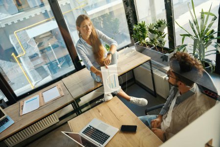 Foto de High angle view of businesswoman with business graph and her male colleague looking at each other in sunny office. Concept of teamwork - Imagen libre de derechos