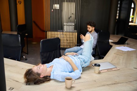 Foto de Businesswoman lying and sleeping on wooden table while her male colleague using smartphone in coworking office. Concept of break and rest at work - Imagen libre de derechos