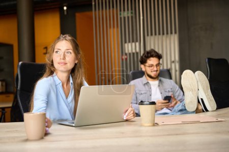 Foto de Businesswoman looking away during work on laptop while her blurred male colleague using smartphone in coworking office. Concept of modern business lifestyle - Imagen libre de derechos